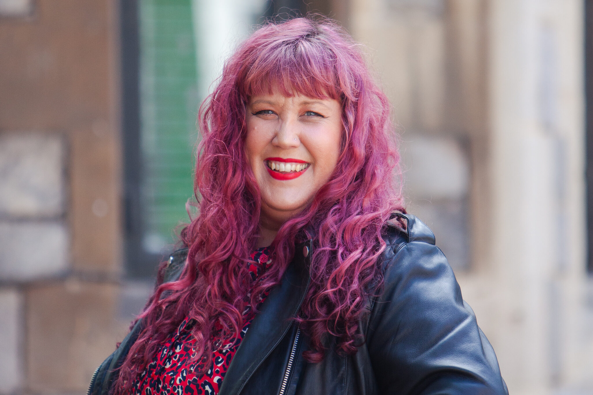 Laura Summers, Marketing Expert with long curly pink hair, wearing a black leather jacket and patterned top, smiling in an urban setting.